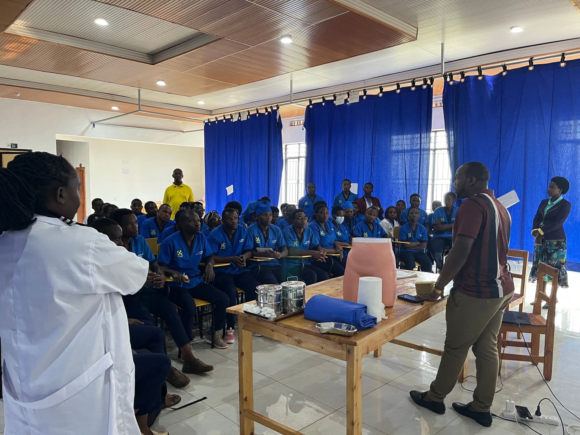 A facilitator leading a practical session for a room of students in blue overalls seated around a demonstration table.
