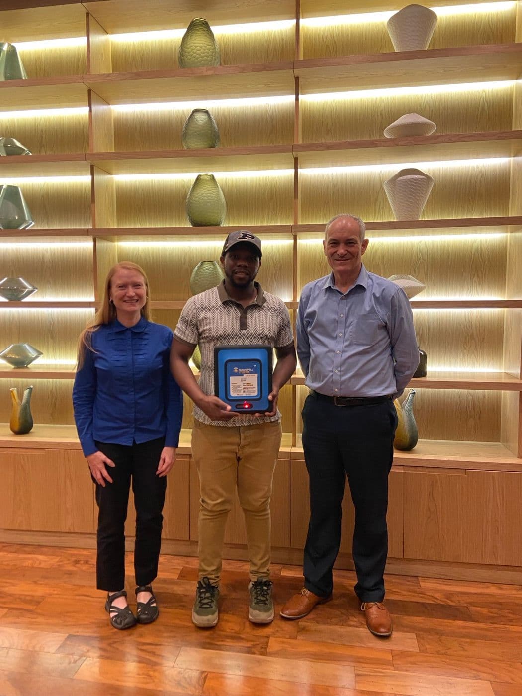 Three people standing together indoors, one holding a framed plaque marking a new partnership.